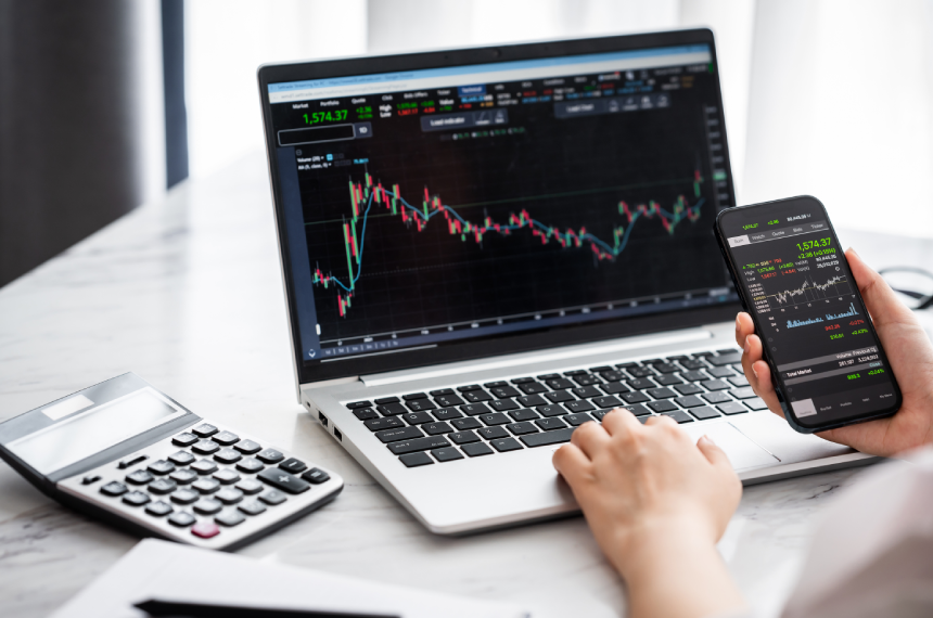 Person analyzing stock market charts on a laptop and smartphone, with a calculator nearby on a desk.