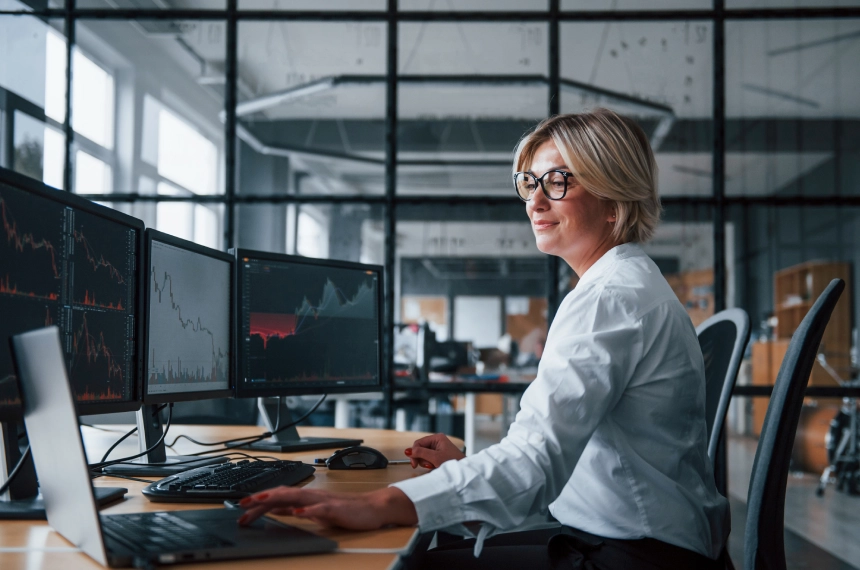 A person in a white shirt works at a desk with multiple monitors displaying graphs in a modern office setting.