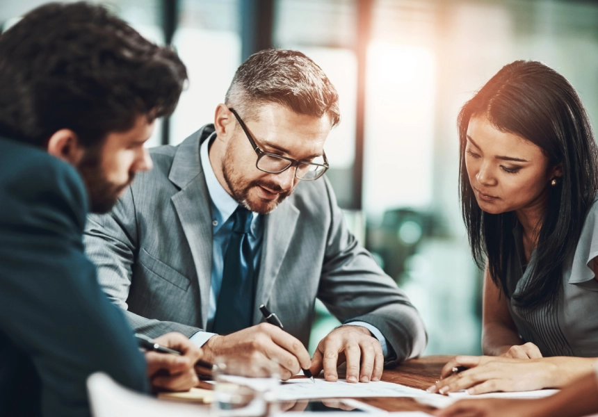 Three people in business attire sit around a table, discussing documents. One person is writing while the others observe.