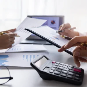 Three hands work with papers, charts, and a calculator on a desk as the Advanced laptop sits in the background.