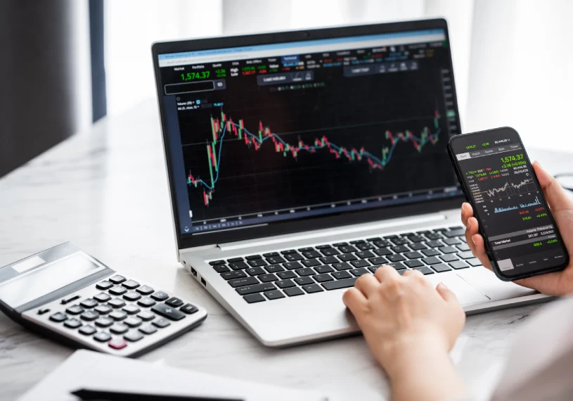 Person analyzing stock market charts on a laptop and smartphone, with a calculator nearby on a desk.