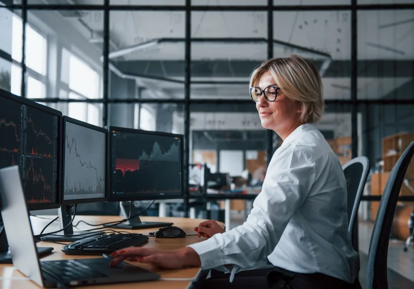 A person in a white shirt works at a desk with multiple monitors displaying graphs in a modern office setting.